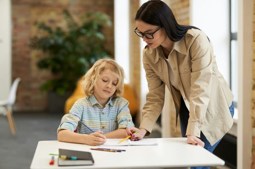 Supportive young female teacher in glasses helping little boy with the task